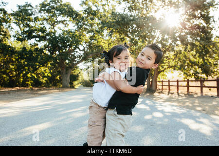 Portrait of brother carrying cute sister while standing on road against trees in park Stock Photo