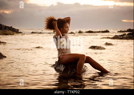 Teenage girl sitting on rock with hand in hair amidst sea Stock Photo