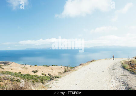 Distant view of hiker standing on mountain by sea against sky during sunny day Stock Photo