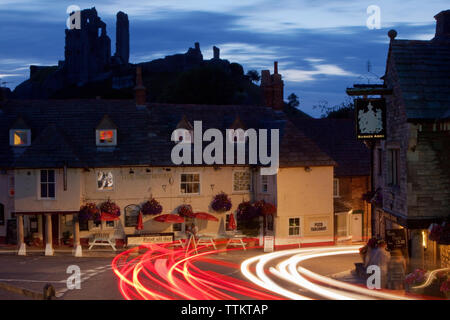 Traffic light trails in a long exposure photograph taken as cars pass The Bankes Arms and The Greyhound pub in front of the ruins of Corfe Castle Stock Photo