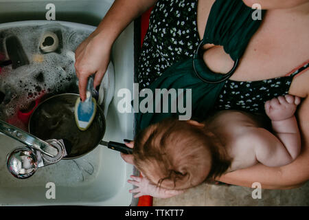 Mother carrying newborn child does dishes at home in kitchen Stock Photo