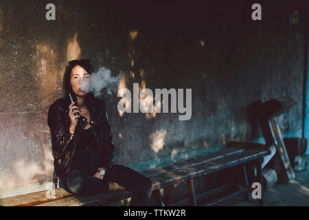 Portrait of confident young woman smoking electronic cigarette while sitting on bench against wall on footpath Stock Photo