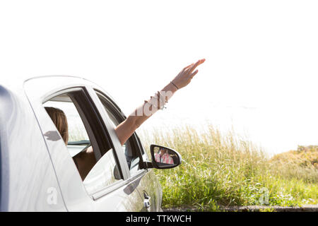 Woman waving hand through window while traveling in car against clear sky during sunny day Stock Photo