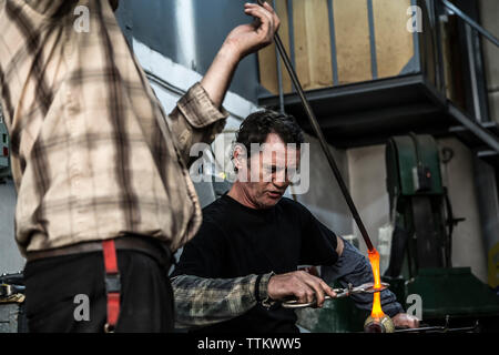Glass blower cutting molten glass with scissors in workshop with coworker Stock Photo