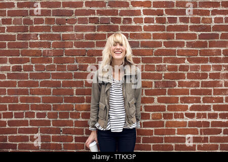 Portrait of cheerful young woman holding disposable glass against brick wall Stock Photo