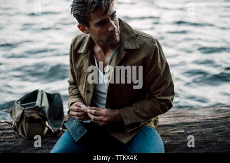 Man holding cigarette looking away while sitting by river on log Stock Photo