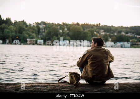 Rear view of man smoking while sitting on log by river against sky Stock Photo