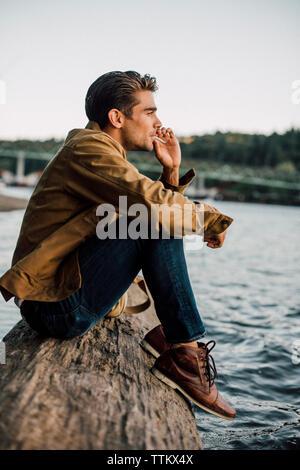 Man smoking while sitting on log by river against clear sky Stock Photo