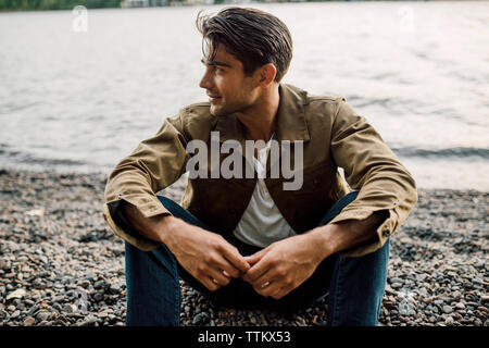 Thoughtful man looking away while sitting at riverbank during sunset Stock Photo