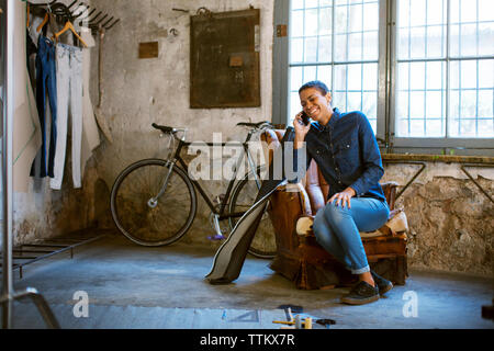 Smiling woman talking on phone while sitting in old workshop Stock Photo
