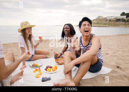 Cheerful friends eating fruits while sitting on beach Stock Photo