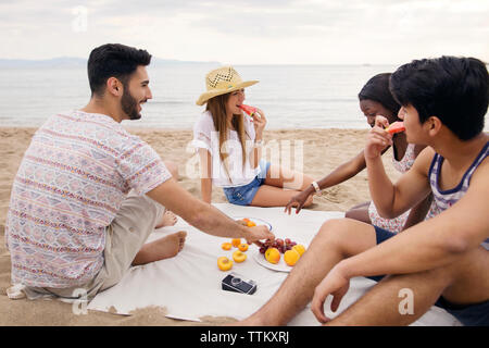 Happy friends eating fruits while sitting on beach Stock Photo