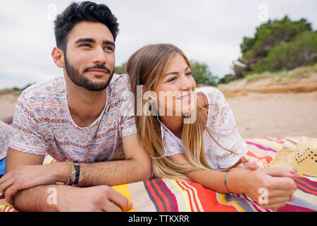 Thoughtful couple looking away while lying on blanket at beach Stock Photo