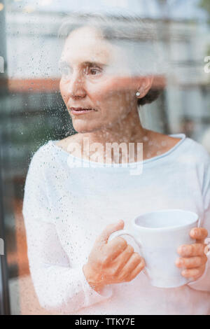 Thoughtful mature woman looking through window while holding coffee mug at home Stock Photo