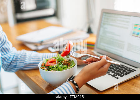 Cropped image of woman holding fork with tomato slice by laptop computer at wooden table Stock Photo