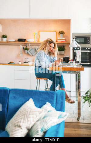 Full length of woman using smart phone while sitting at table in kitchen Stock Photo