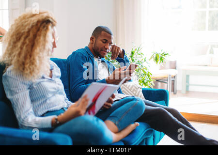 Woman reading magazine while man using smart phone on sofa at home Stock Photo