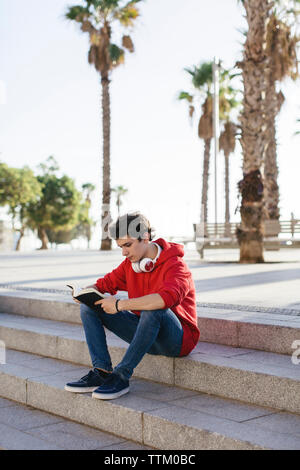 Serious teenage boy reading book while sitting on steps in city Stock Photo
