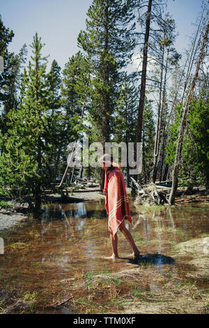 Woman wrapped in blanket walking on wet field against trees at Yellowstone National Park Stock Photo