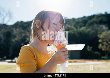 Cute girl eating ice cream cone while standing against sky at park during sunny day Stock Photo