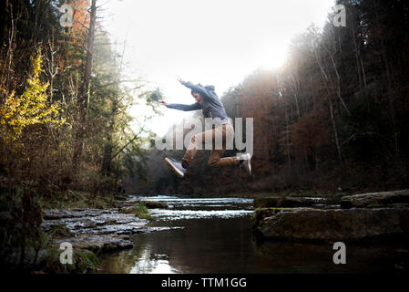 Side view of teenage boy jumping over river against trees in forest Stock Photo