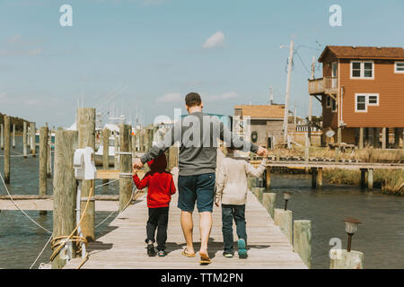 Rear view of father with sons walking on pier over sea against sky during sunny day Stock Photo