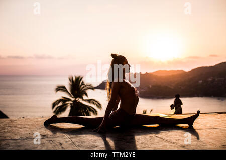 Woman practicing yoga on retaining wall against sea and sky during sunset Stock Photo