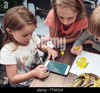 High angle view of sisters using mobile phone while brother drawing on table at home Stock Photo