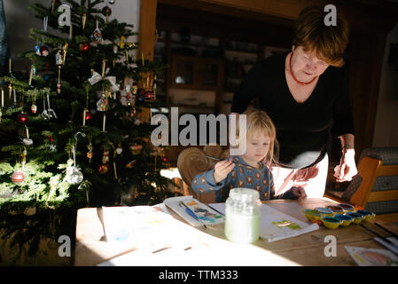 Grandmother looking at granddaughter painting on book in house during christmas Stock Photo