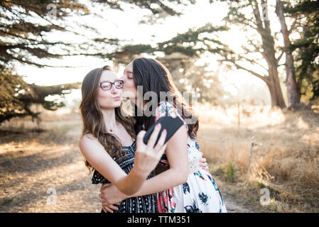 Happy young woman kissing sister talking selfie with smart phone while standing against trees in forest Stock Photo