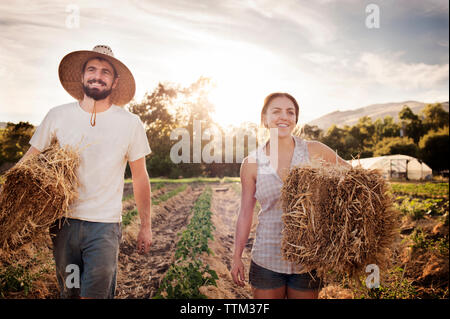 Happy thoughtful farmers carrying hay bales while walking on field Stock Photo