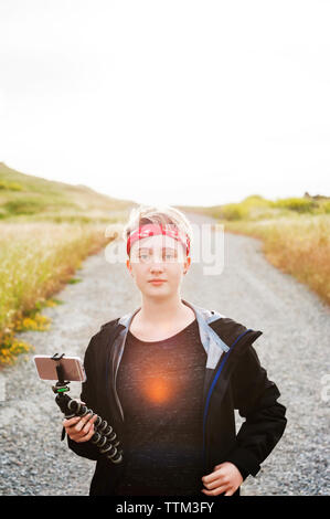 Portrait of teenage girl holding smart phone in tripod on country road Stock Photo