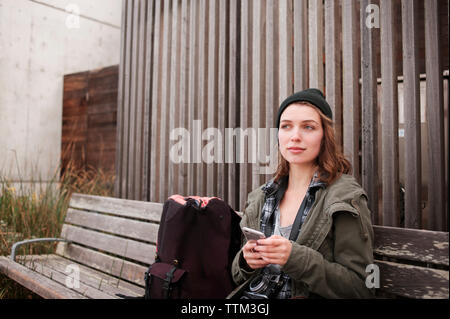 Thoughtful female hiker holding smart phone while sitting on bench Stock Photo