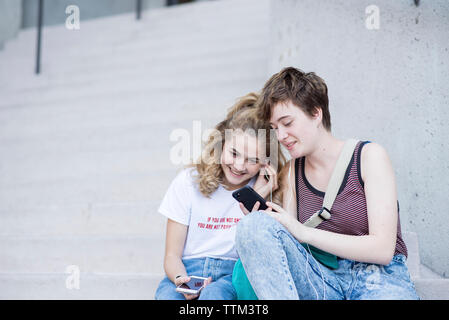 Female friends listening music while looking at smart phone on steps in city Stock Photo
