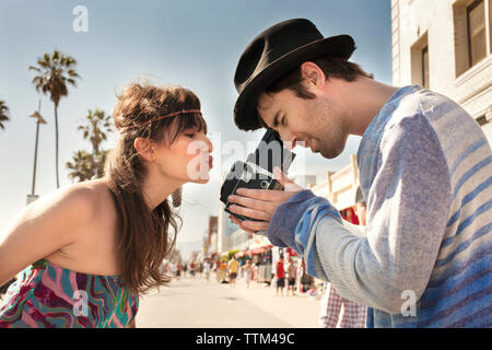 Side view of woman puckering lips while man photographing through vintage camera on sidewalk Stock Photo