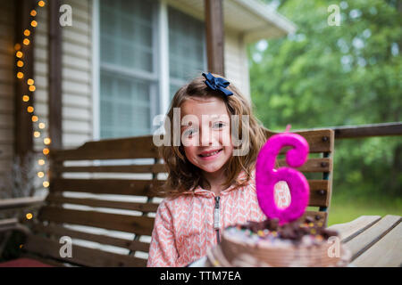 Portrait of happy girl sitting by birthday cake with number 6 candle at porch Stock Photo