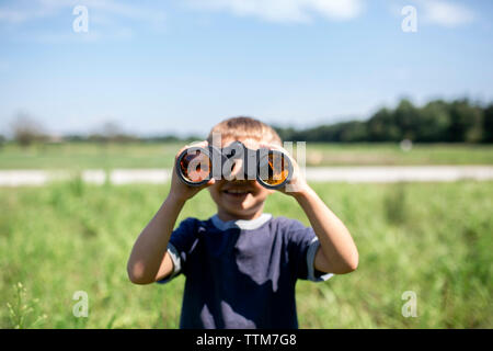 Playful boy looking through binoculars while standing on field during sunny day Stock Photo