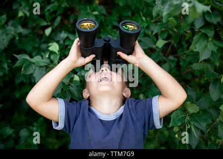 Playful boy looking through binoculars while standing against plants Stock Photo