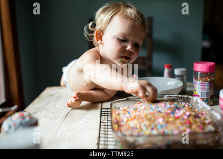 Toddler girl sneaking colorful sprinkles off of freshly baked cake Stock Photo