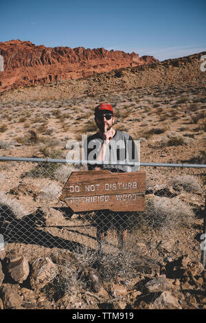 Man gesturing silence while standing by information sign on fence at Valley of Fire State Park during sunny day Stock Photo