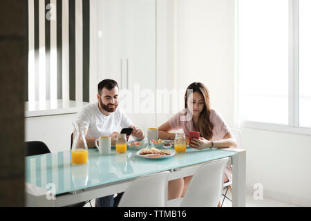 Young couple using mobile phones while having breakfast at dining table in rental house Stock Photo