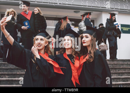 Happy female friends wearing graduation gowns taking selfie while standing on steps Stock Photo