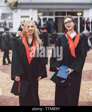 Portrait of happy female students wearing graduation gowns while standing against building Stock Photo