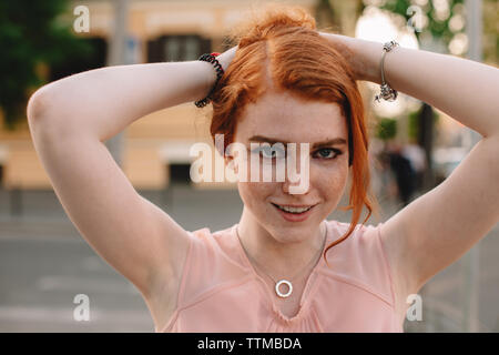 Portrait of young redheaded woman holding hair while standing in city