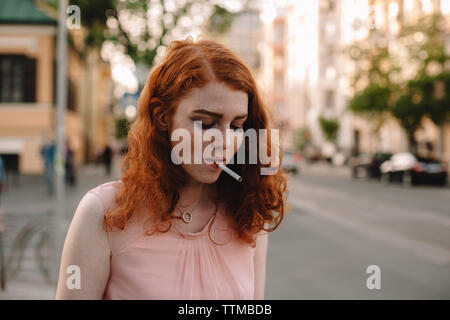 Young redheaded woman with cigarette standing on street in city Stock Photo