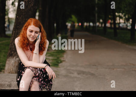 Young redhead woman talking on phone while sitting in park Stock Photo