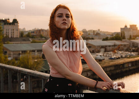 Portrait of young redheaded woman standing on bridge at sunset