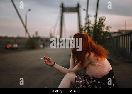 Young redheaded woman smoking cigarette while sitting on bridge Stock Photo
