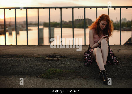 Young redhead woman using mobile phone while sitting on bridge in city Stock Photo