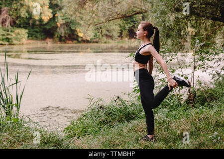 Side view of sporty woman stretching by lake in park Stock Photo